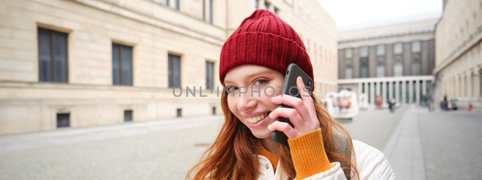 Mobile broadband and people. Smiling young redhead woman walks in town and talks on mobile phone, calling friend on smartphone, using internet to make a call abroad.