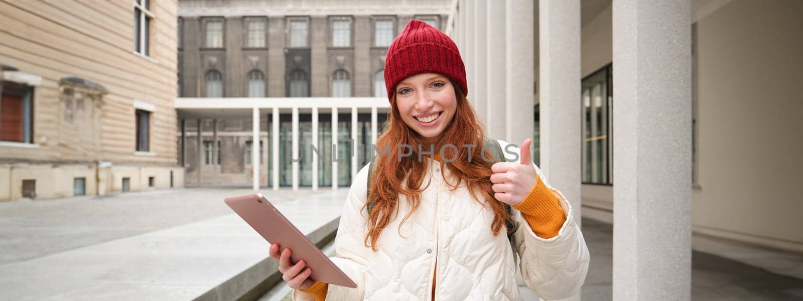 Stylish ginger girl, tourist walks with digital tablet around city, woman connects to iternet on her gadget, looking up information, texting message by Benzoix