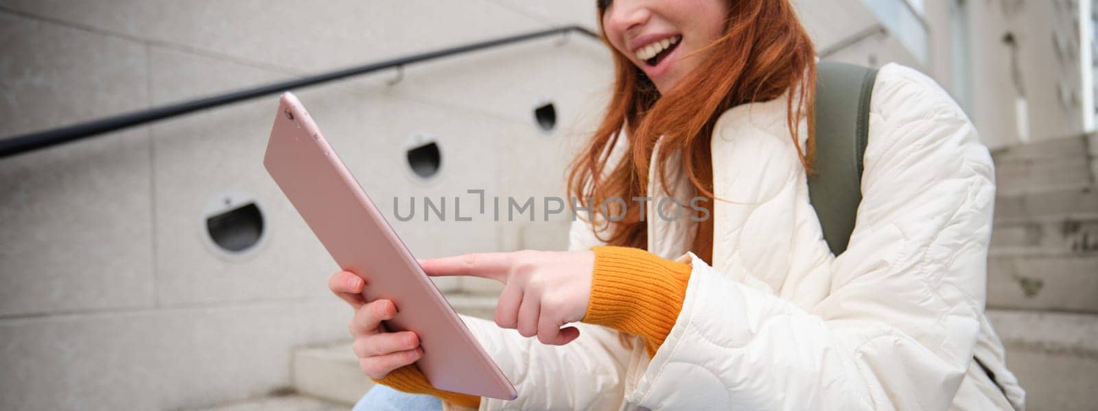 Young stylish girl, redhead female students sits on stairs outdoors with digital tablet, reads, uses social media app on gadget, plays games while waits on street by Benzoix