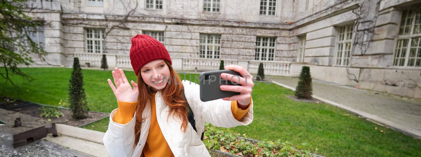 Portrait of girl blogger records video and says hi at phone camera. Young woman takes selfie with smartphone app, waves hand, video chats in application.