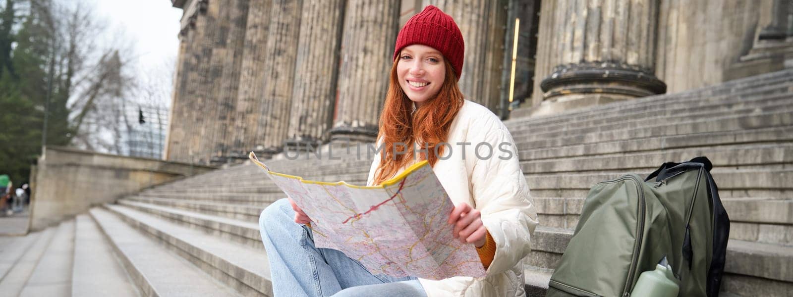 Tourism and lifestyle concept. Young redhead woman looking at city map, plans a route for sightseeing day, sits outdoors on stairs and rests.