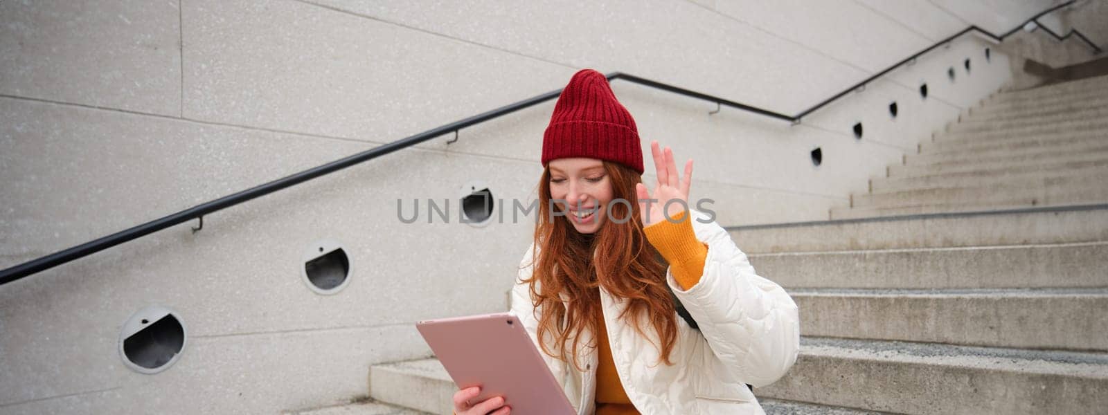 Joyful redhead teen girl, student with digital tablet, says hello, waves hand at gadget camera, connects to video chat, talks to friend in application.