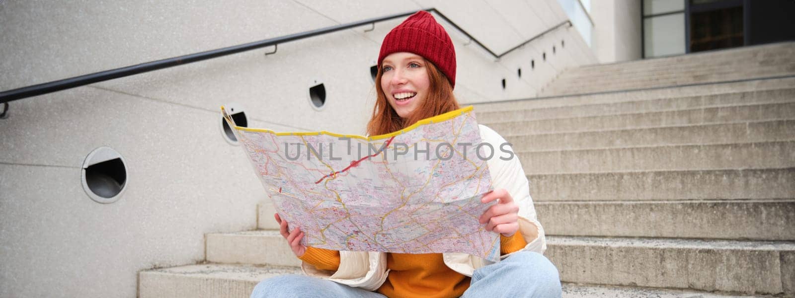 Young smiling redhead girl, tourist sits on stairs outdoors with city paper map, looking for direction, traveller backpacker explores city and looks for sightseeing.