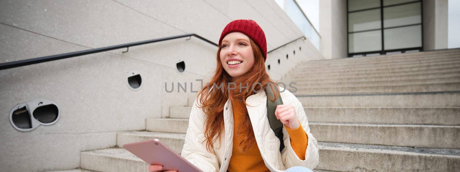 Happy redhead woman in red hat, sits on stairs outdoors, uses tablet, connects to wifi near public building, reads digital book while waits on street.