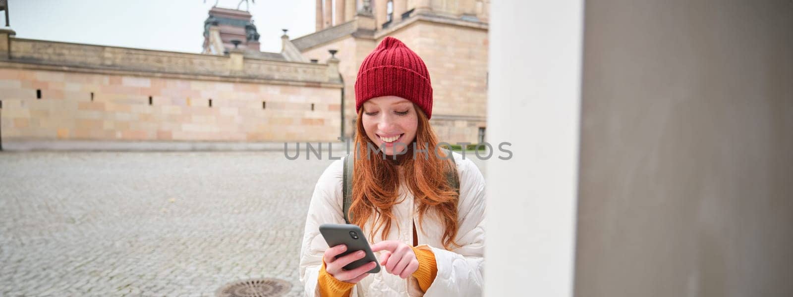 Smiling redhead girl tourist, walks around city and explores popular landmarks, sightseeing, holding smartphone, looking at her mobile app and checks with city map by Benzoix