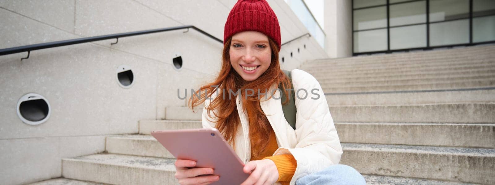 Young beautiful girl with long red hair, smiles, uses internet application, connect to wifi near campus, sits on stairs with digital tablet by Benzoix
