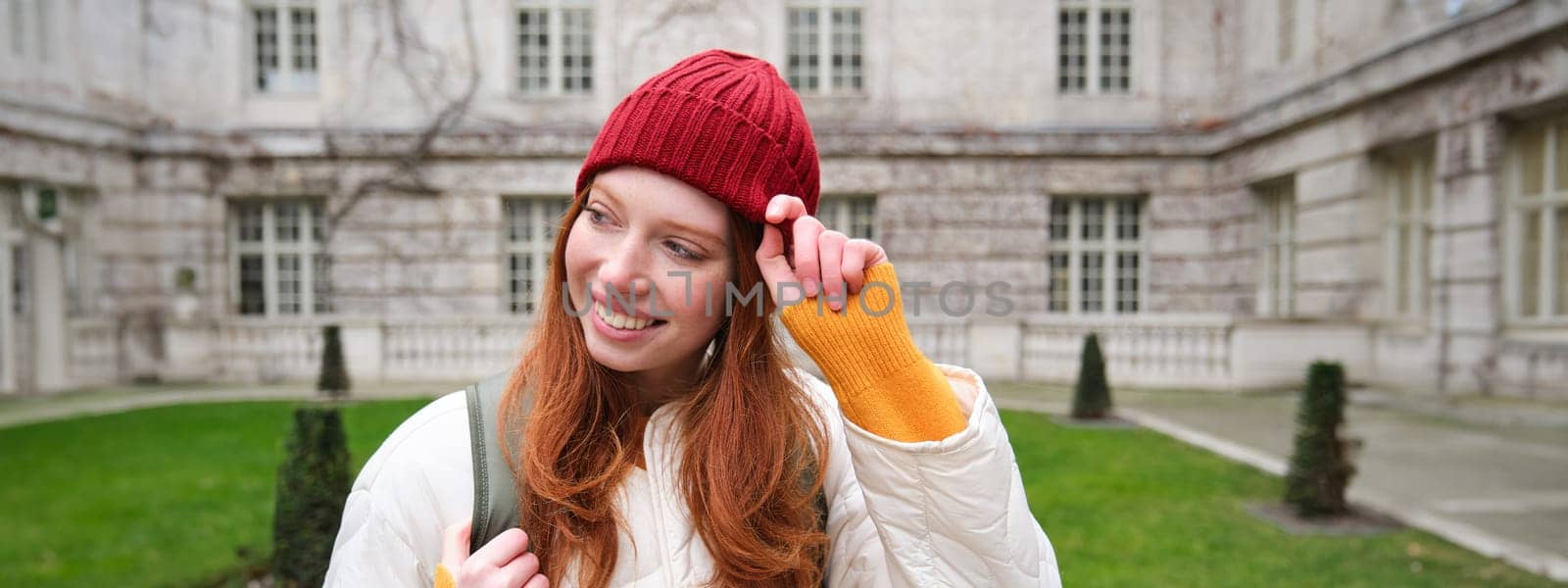 Beautiful redhead woman with backpack, tourist walking around city park and looks around, wears red hat and warm coat.