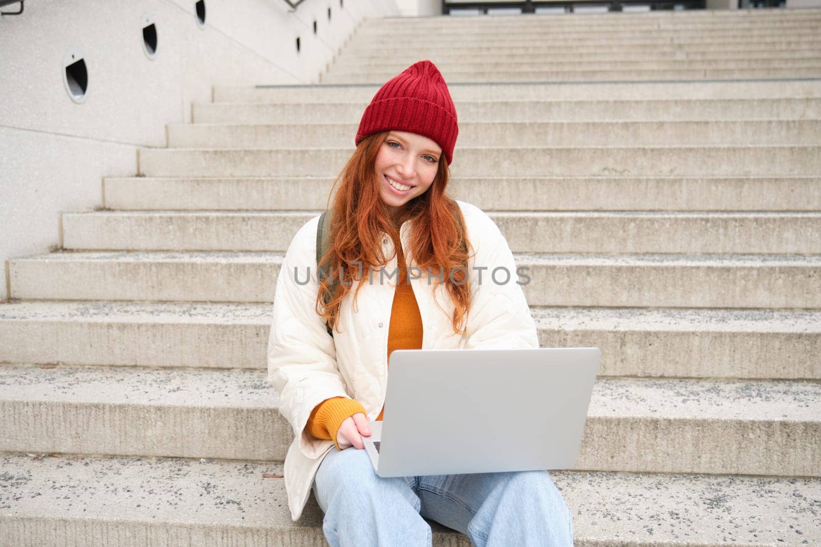 Smiling redhead girl, student sits on stairs outdoors and uses laptop, connects to public wifi in city and works on project, uses internet on computer.