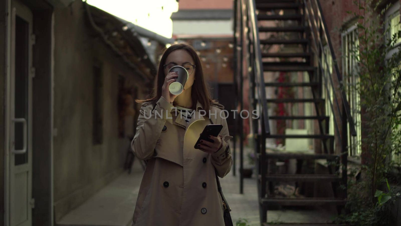 A young woman walks along a narrow street late at night and chats on her smartphone. A girl with glasses walks with a phone in her hands and drinks coffee. 4k