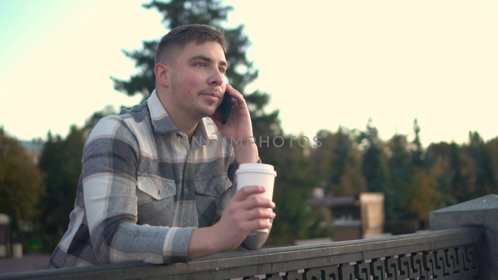 A young man speaks on the phone with a glass of coffee in his hand. A man with a phone near the fence on the embankment with a hot drink in a glass. 4k