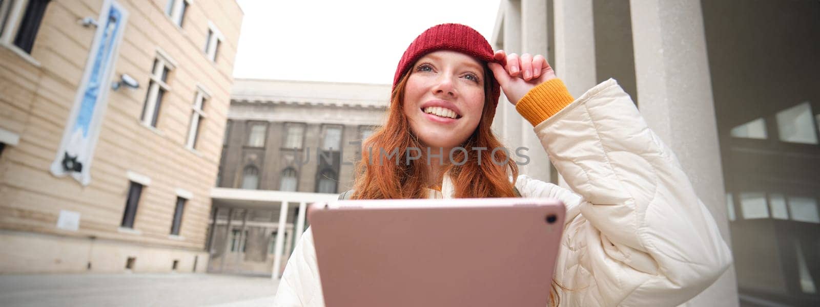 Happy redhead girl in red hat, walks around city with digital tablet, connects to public internet wifi and looks for route, looks at map on her gadget by Benzoix