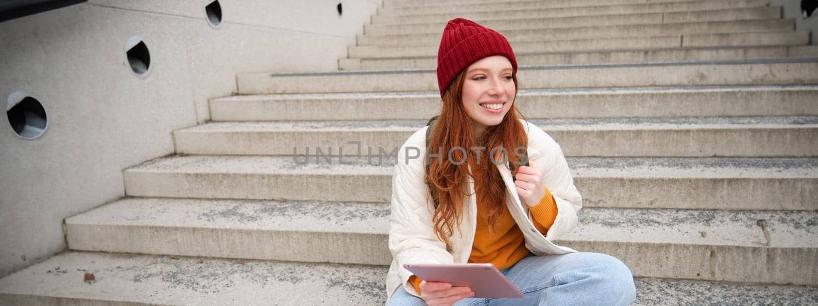 Young beautiful girl with long red hair, smiles, uses internet application, connect to wifi near campus, sits on stairs with digital tablet by Benzoix