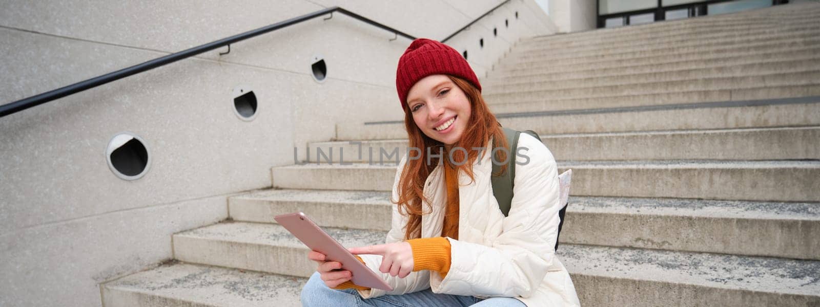 Happy stylish redhead girl, student in red hat, holds digital tablet, uses social media app, searches something online, connects to wifi.