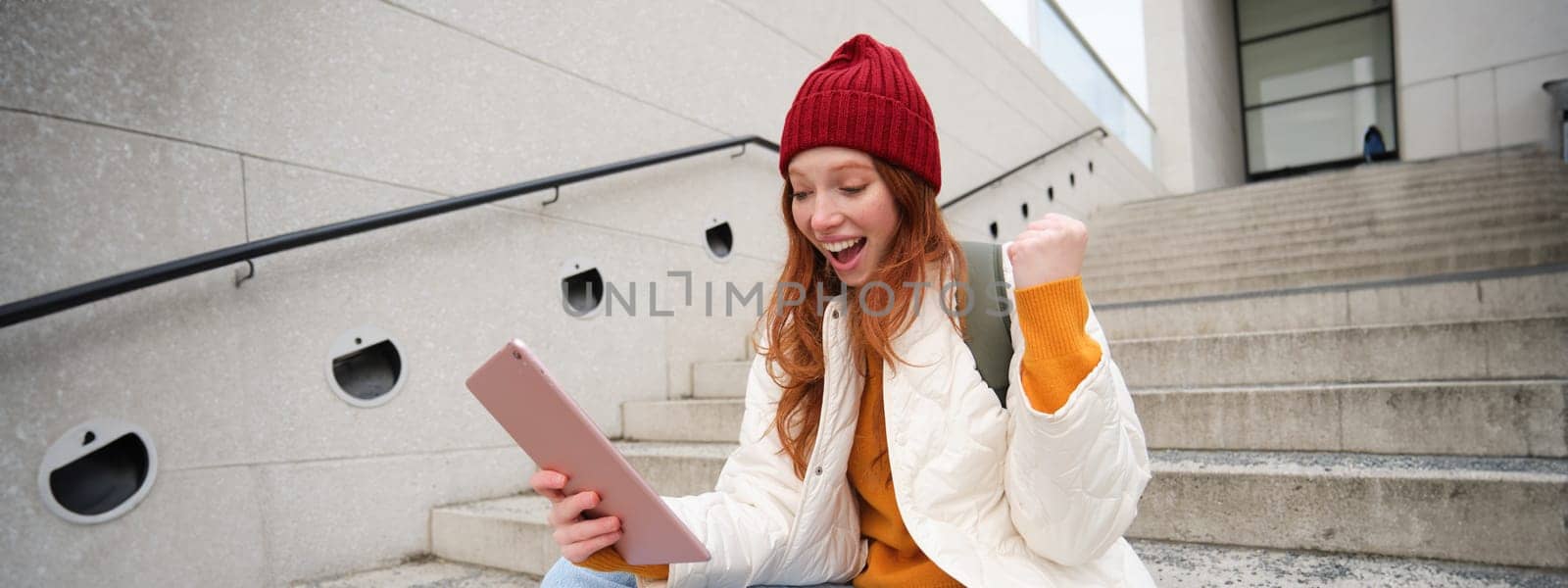 Happy stylish redhead girl, student in red hat, holds digital tablet, uses social media app, searches something online, connects to wifi by Benzoix