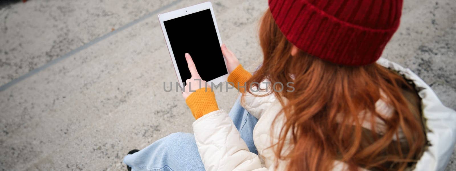 Rear view of redhead girl touches digital tablet screen, touchpad, texts message, uses internet application on gadget, sits on stairs outdoors.