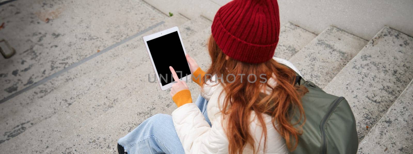 Rear view of redhead girl touches digital tablet screen, touchpad, texts message, uses internet application on gadget, sits on stairs outdoors.