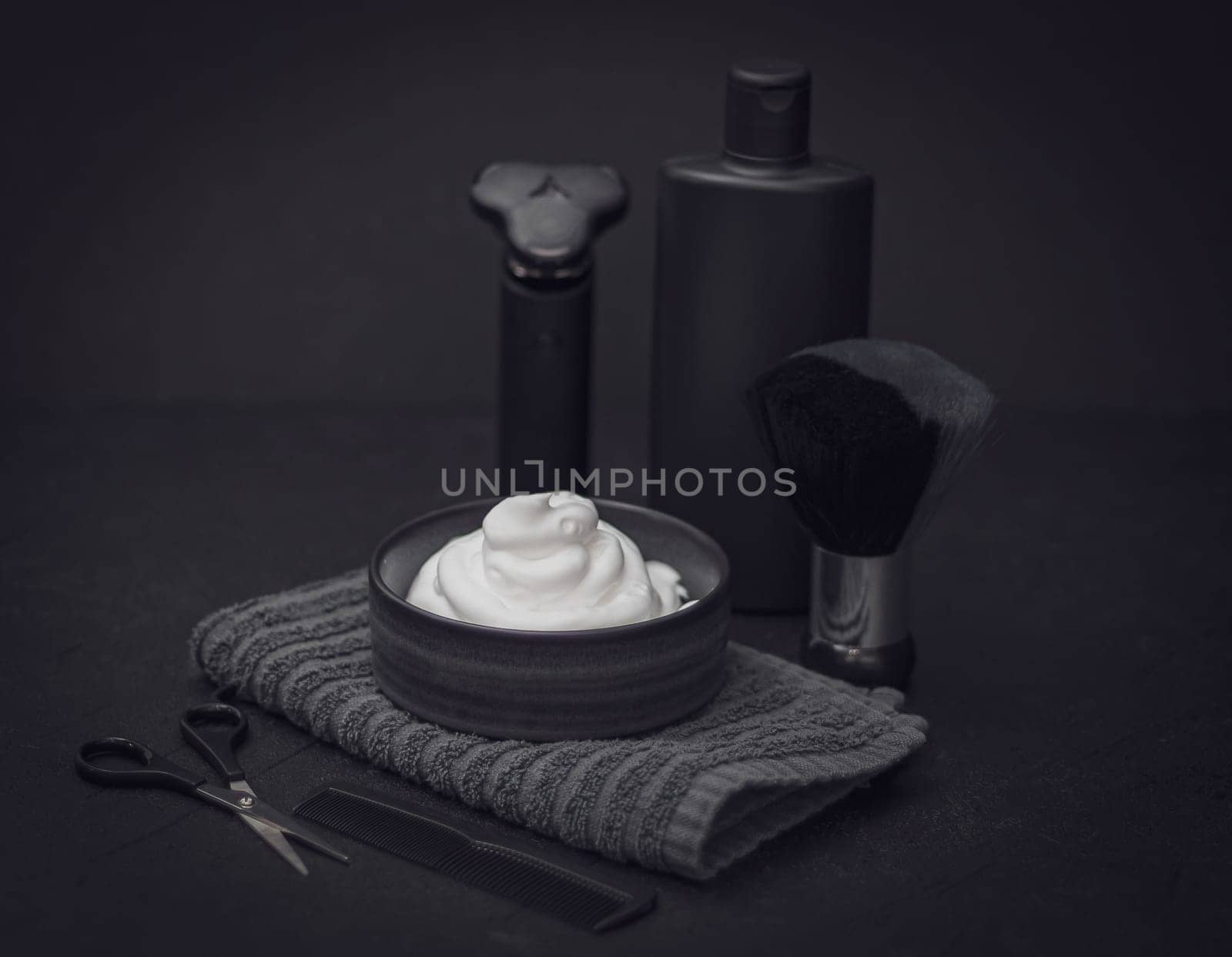 Shaving foam in a bowl with a terry towel, a comb, scissors, a shaving brush, an electric razor and a bottle of aftershave cream on a black background, close-up side view. The concept of a male hairdresser, a beauty salon, a beard shave.
