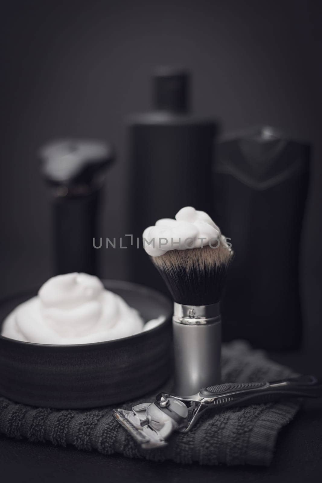 Shaving foam in a bowl with a terry towel, a razor, a shaving brush, an electric razor and a bottle of aftershave cream on a black background, close-up side view with selective focus. The concept of a male hairdresser, a beauty salon, a beard shave.