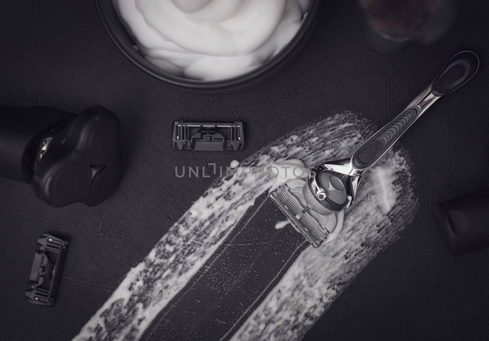 A smear of shaving foam in the center on a black background with a razor, a comb, a bottle of aftershave cream, a bowl of foam and an electric razor with copy space, flat lay close-up. The concept of a male hairdresser, beauty salon, beard shaving.