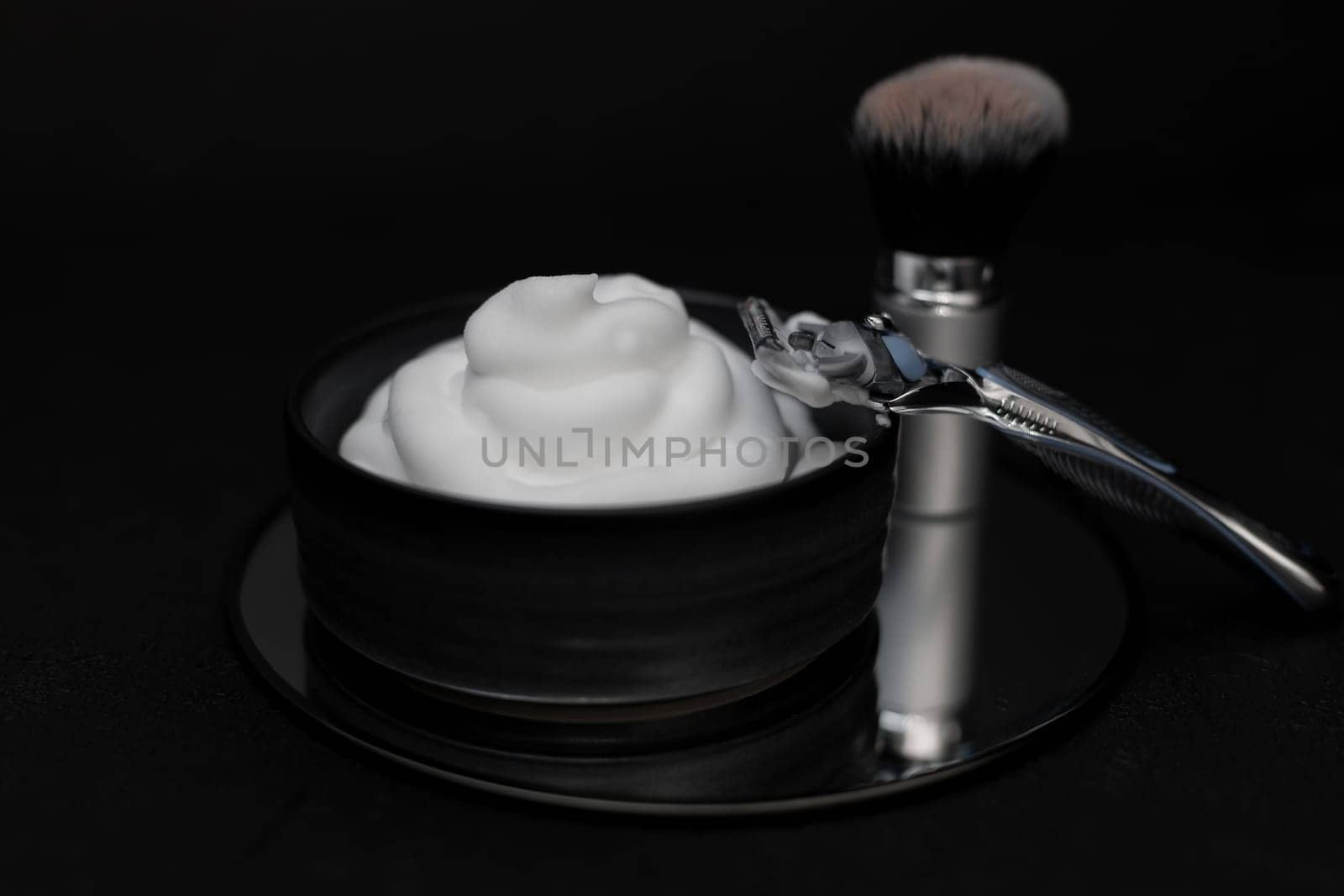 Shaving foam in a bowl, a shaving brush and a razor on a round mirror on a black background, close-up side view. The concept of a male hairdresser, beauty salon, beard shaving.