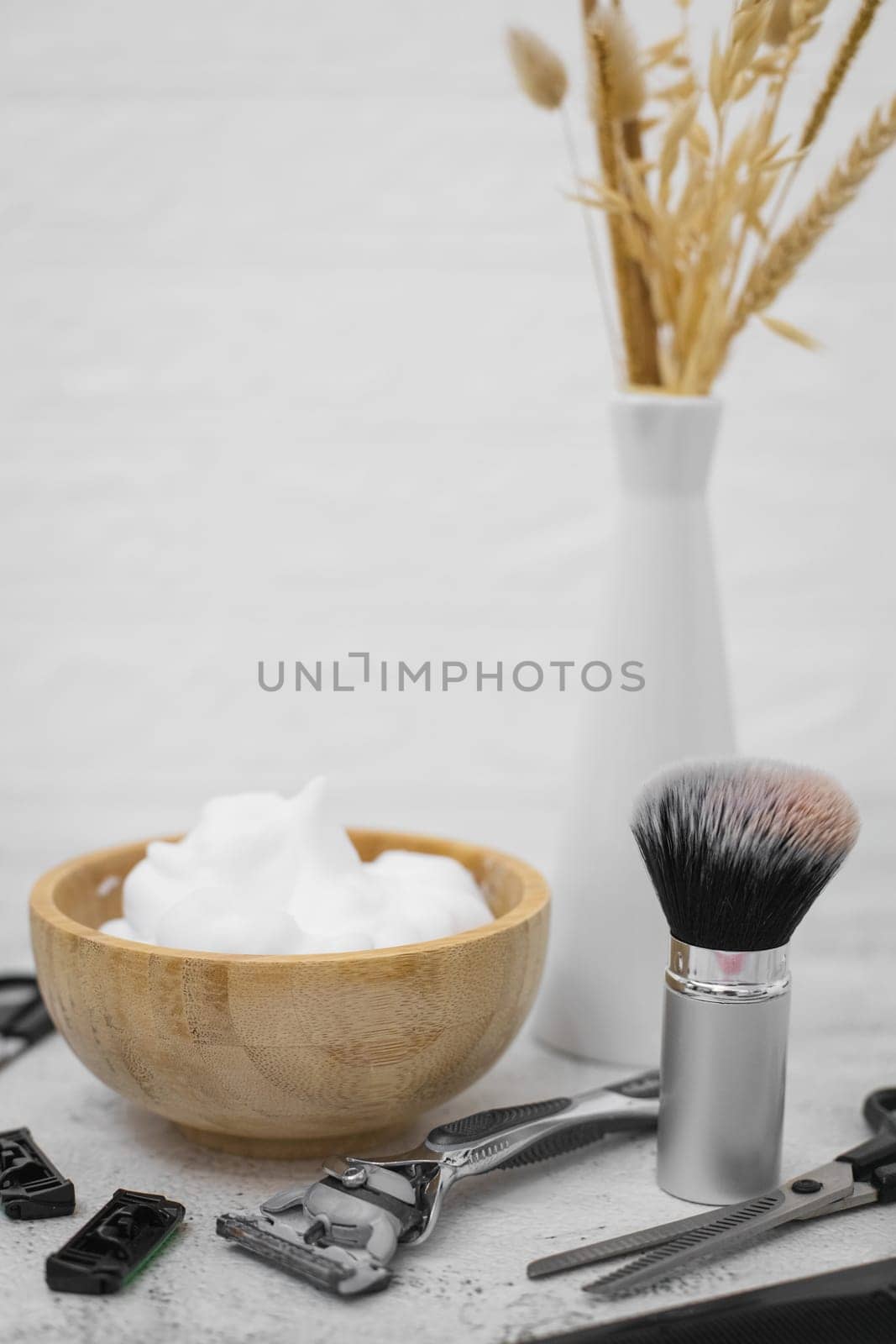 Shaving foam in a wooden bowl, a shaving brush, a razor, scissors, combs, thinning scissors and a vase with dried flowers stand on a light cement background with a white wall, close-up side view. The concept of a male hairdresser, beauty salon, beard shaving.