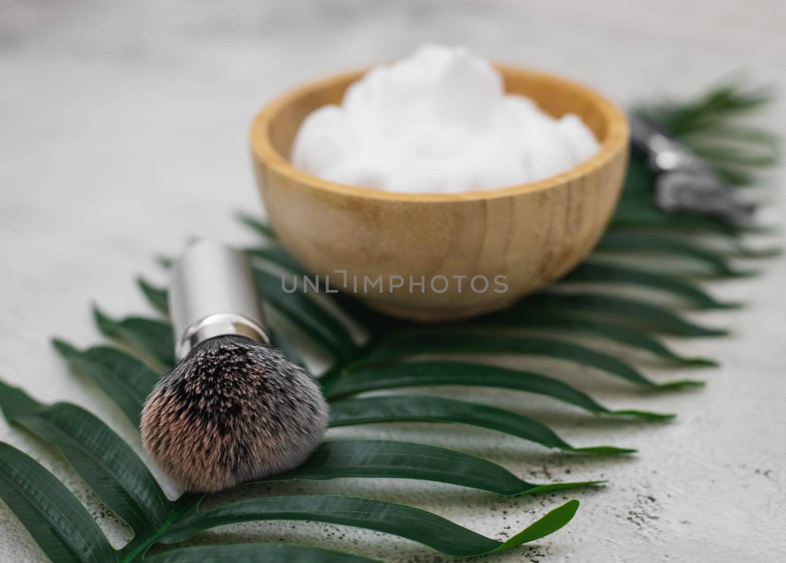 Shaving brush with shaving foam in a wooden bowl on a palm branch lie on a light cement background ,close-up side view with depth of field. The concept of a male hairdresser, beauty salon, beard shaving.