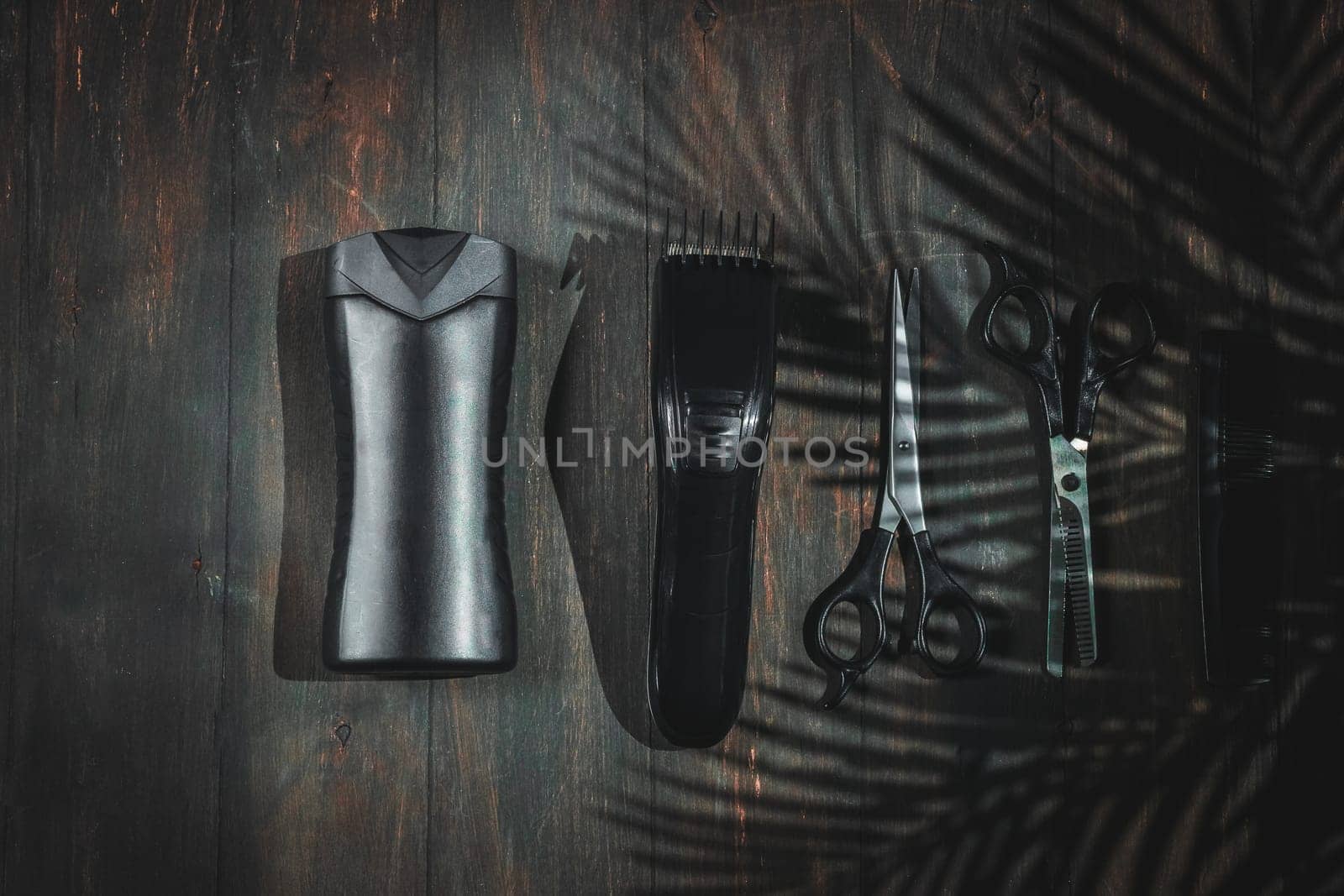 Scissors, combs, shampoo and men's hair clipper lie on a black homemade wooden background with a shadow from a palm branch, flat lay close-up. The concept of a male barbershop.