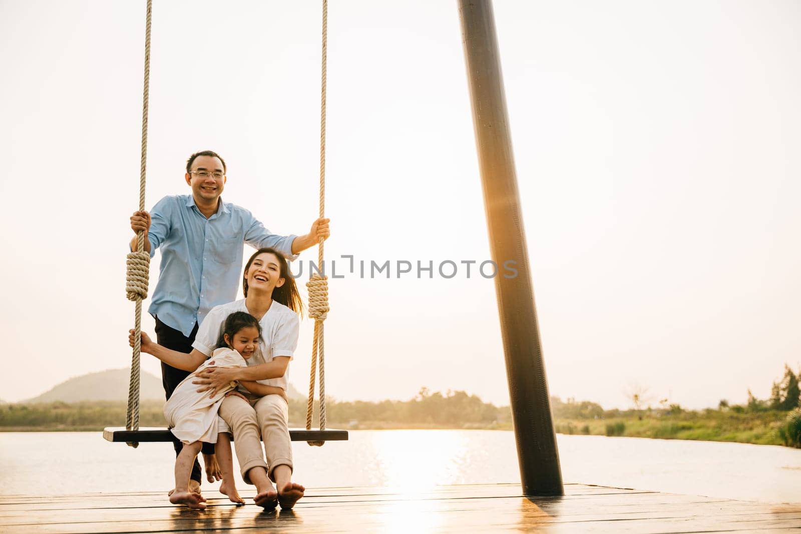 A happy family enjoying a sunny day at the park playground, with the father sitting and pushing the swing while the mother and daughter play together, Happy Family Day