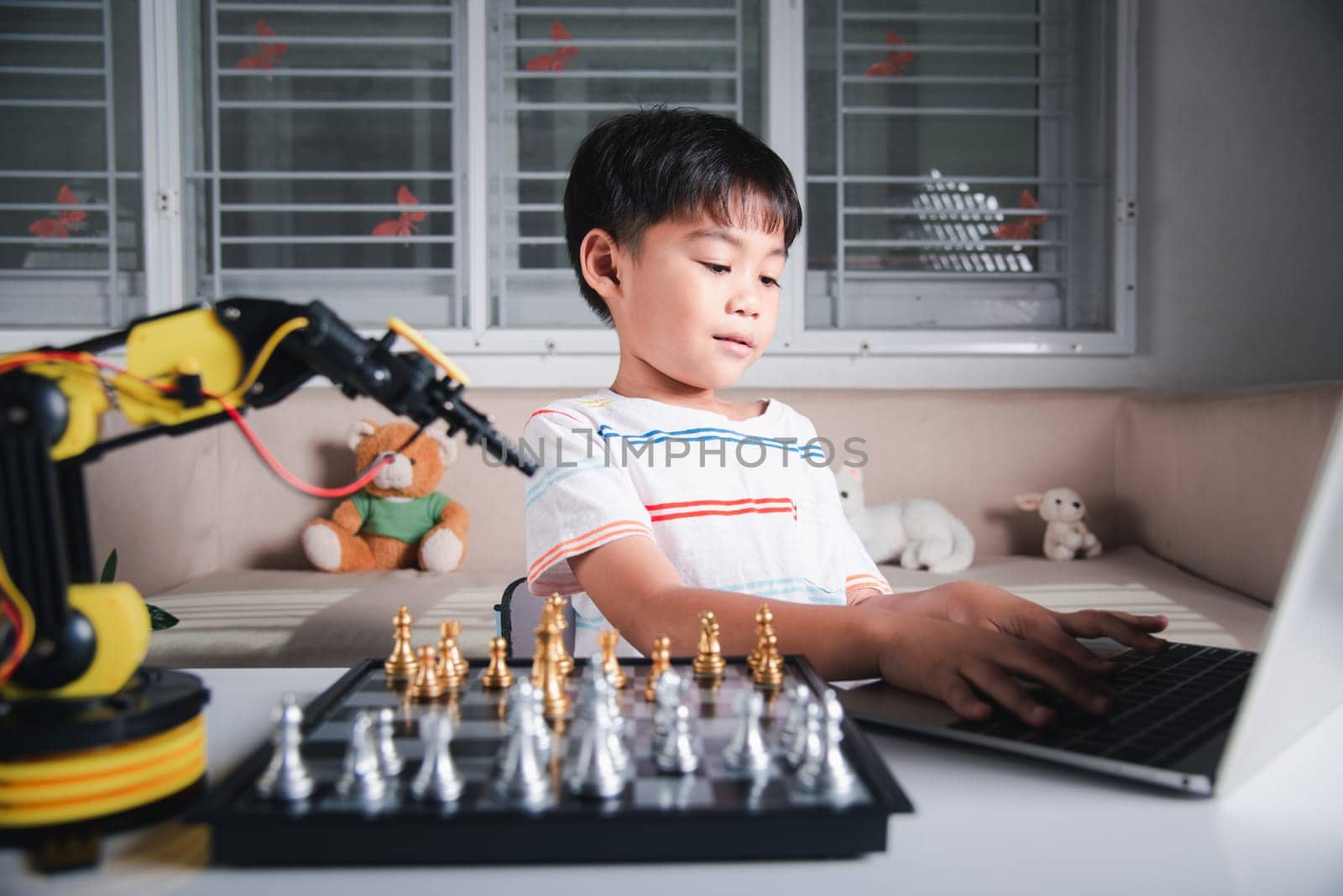 Asian little boy programming code to robot machine arm on laptop for play chess by Sorapop