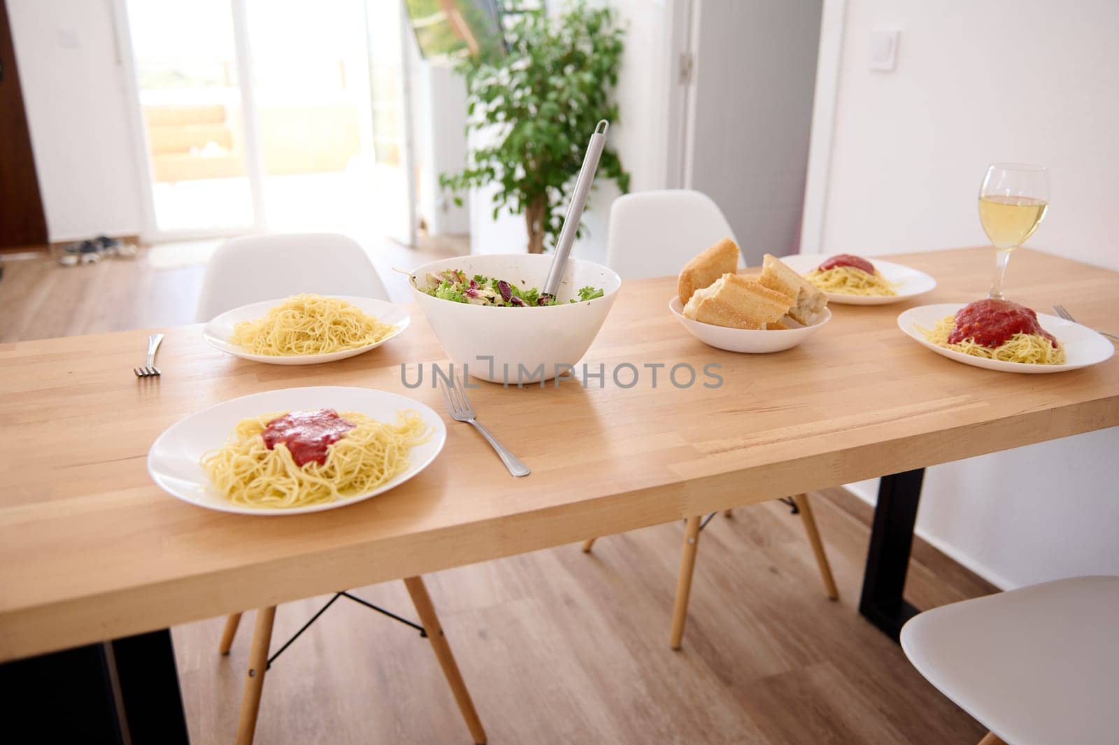 White plates with Italian spaghetti with fresh tomato sauce and a bowl of fresh healthy vegan salad, served on a wooden table in modern interior of a home living room. Traditional Italian cuisine.