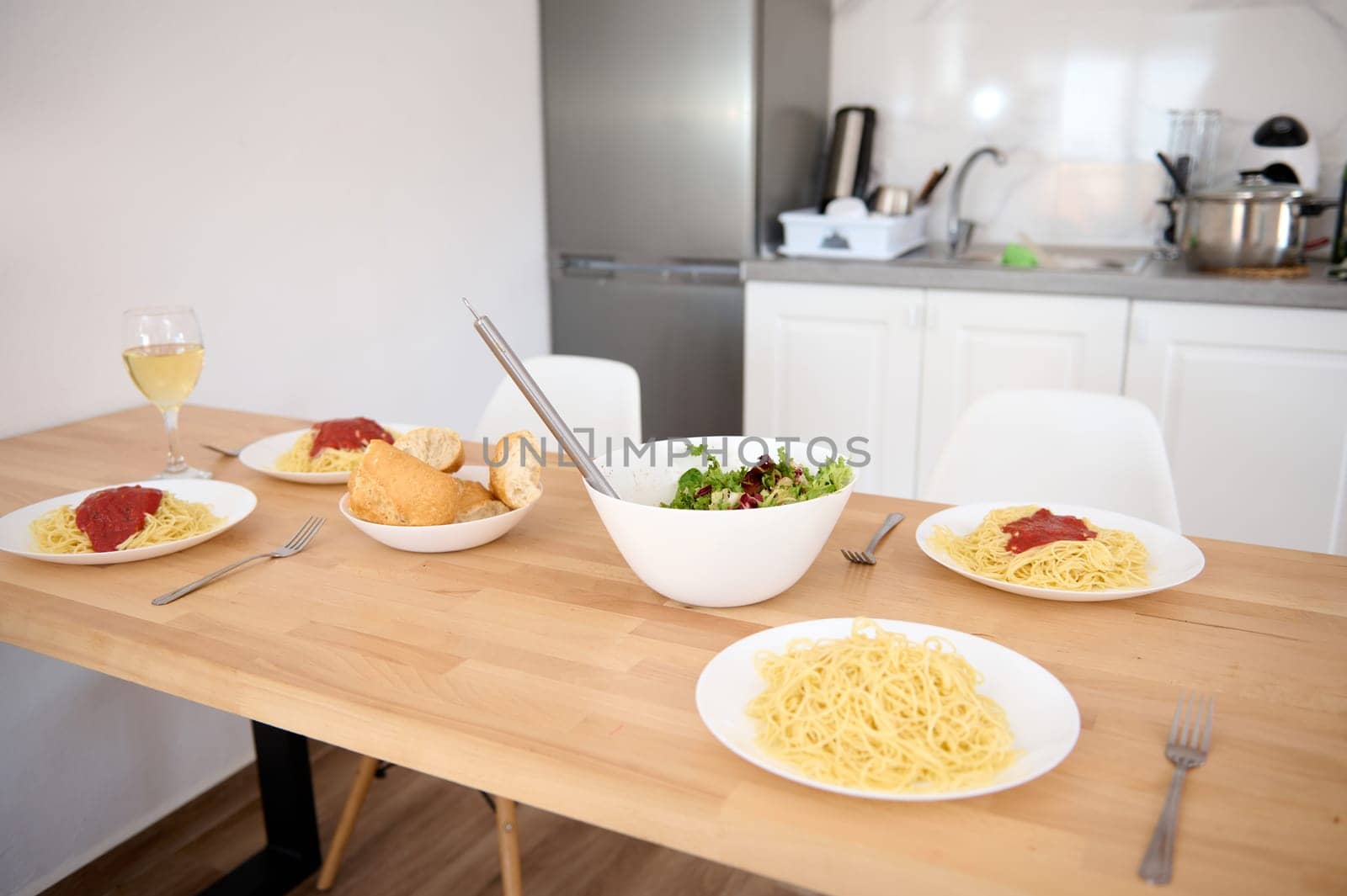 A served table with traditional Italian spaghetti and tomato sauce in white plates, a bowl with wholesome salad and wine in wineglass. Still life with delicious healthy lunch in the home kitchen