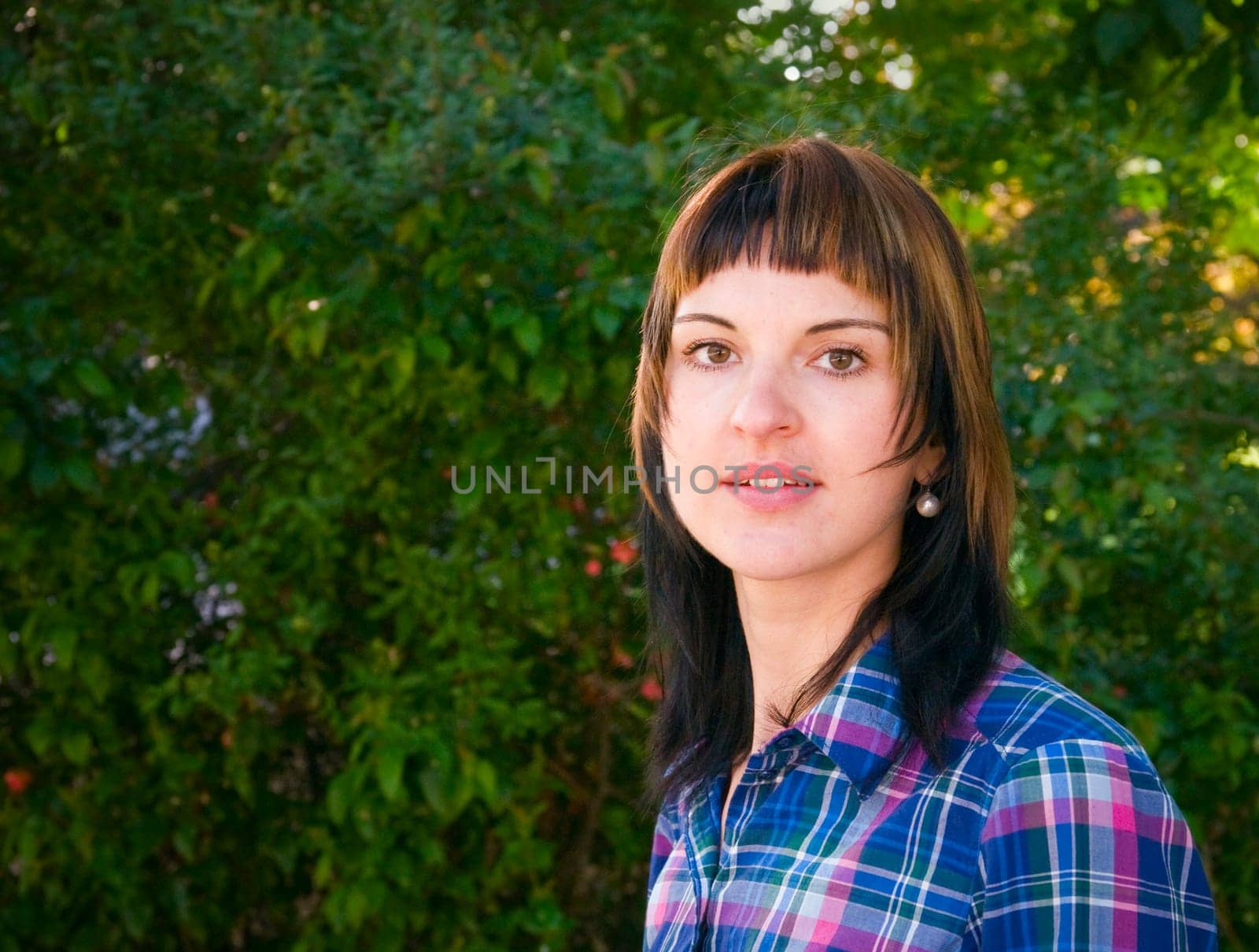 Side head and shoulders portrait of a young brunette woman in front of a green hedge in summer and looking at camera
