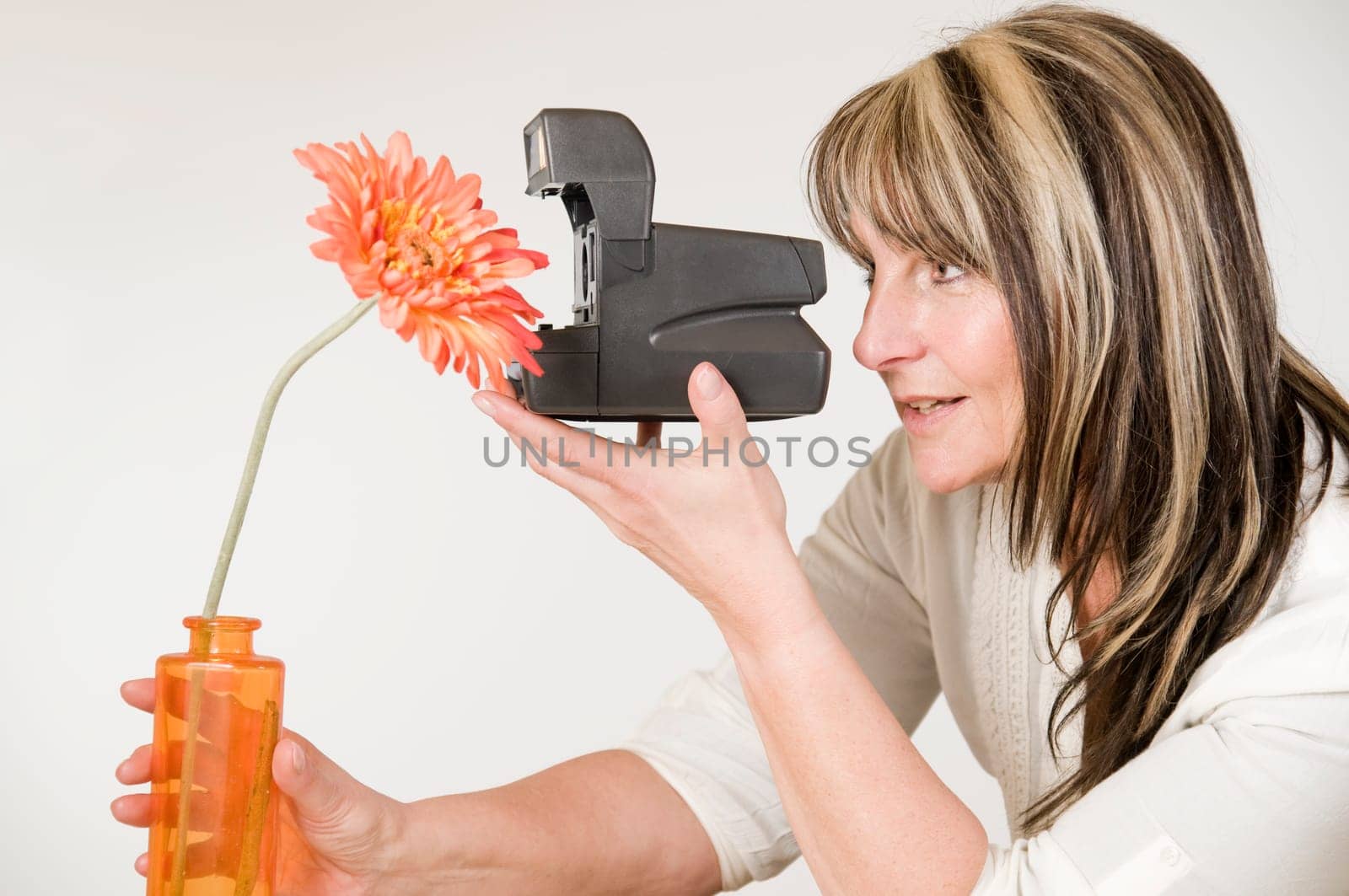 Head and shoulders shot of middle-aged woman with instant camera and orange-colored artificial flower against neutral bright studio background