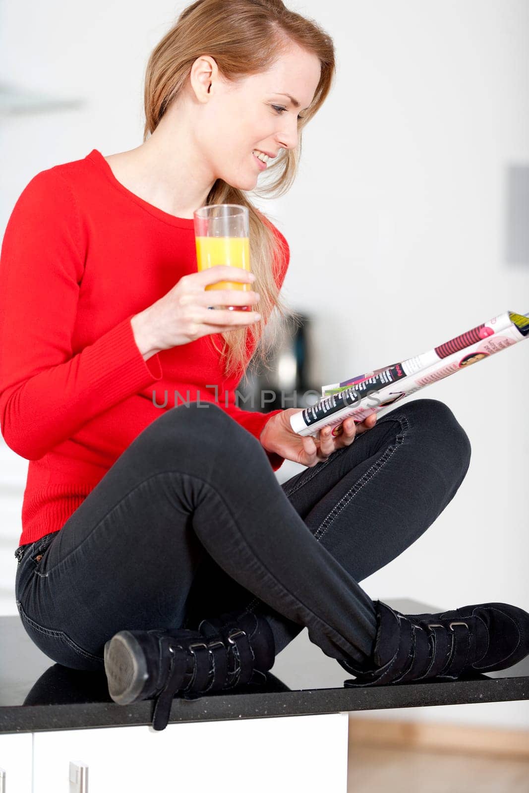 Young woman sat on kitchen work top, enjoying a glass og juice and reading a magazine