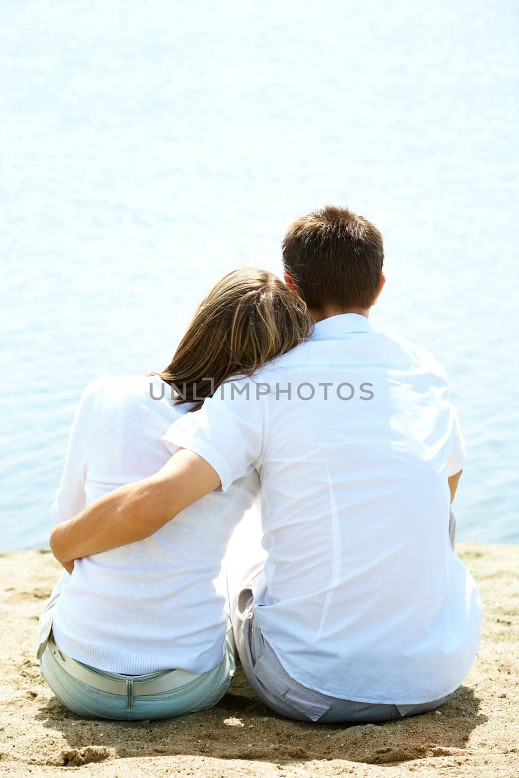 Backs of serene couple sitting on sandy shore in front of blue water