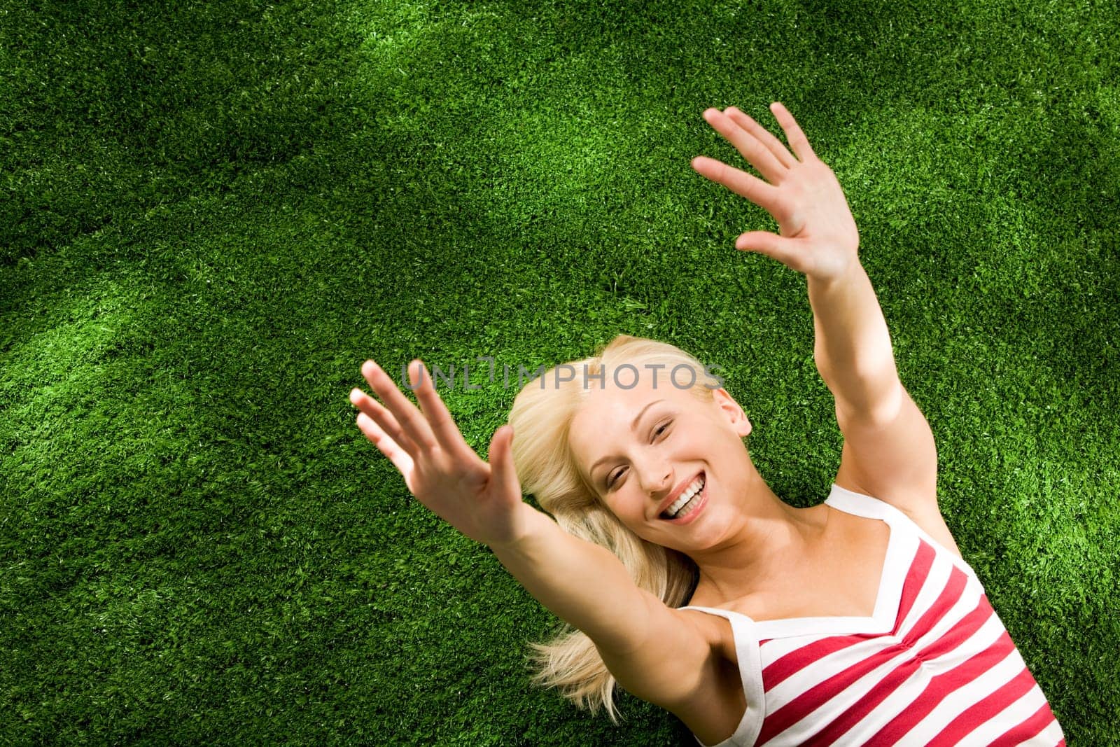 Above view of young glad female lying on green grassland and stretching her arms