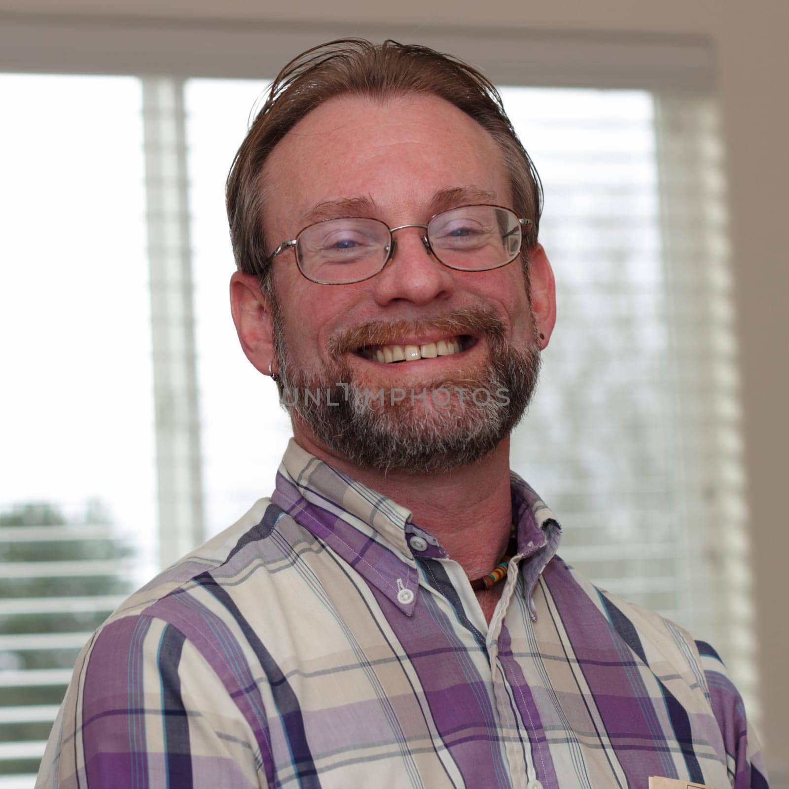 Male wearing eyeglasses in is forties with blonde gray hair and beard smiling in a joyous way showing his upper teeth.