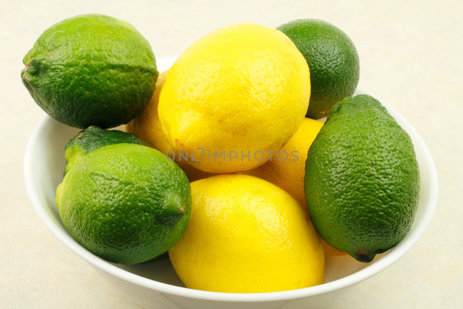 Five limes and four lemons in a white bowl on a tan counter.