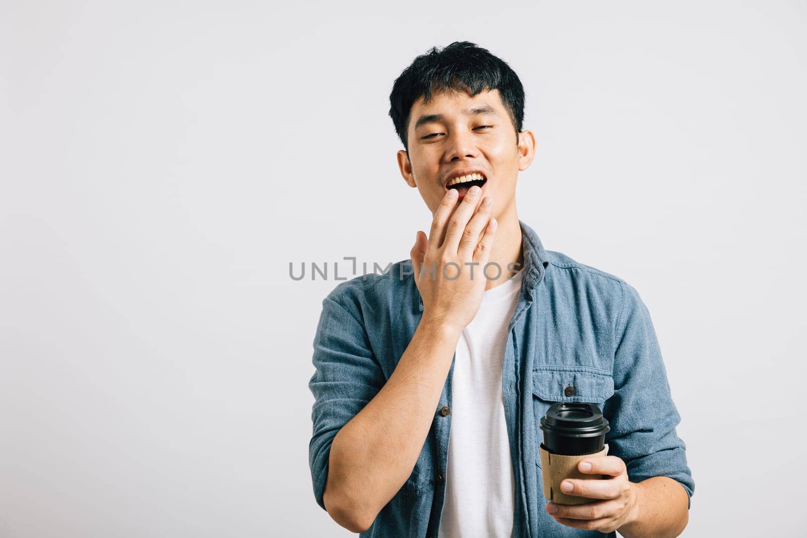 Portrait of an exhausted, overworked man unable to keep his eyes open, clutching a coffee cup. Studio shot isolated on white background, emphasizing his weariness. Copyspace is provided.