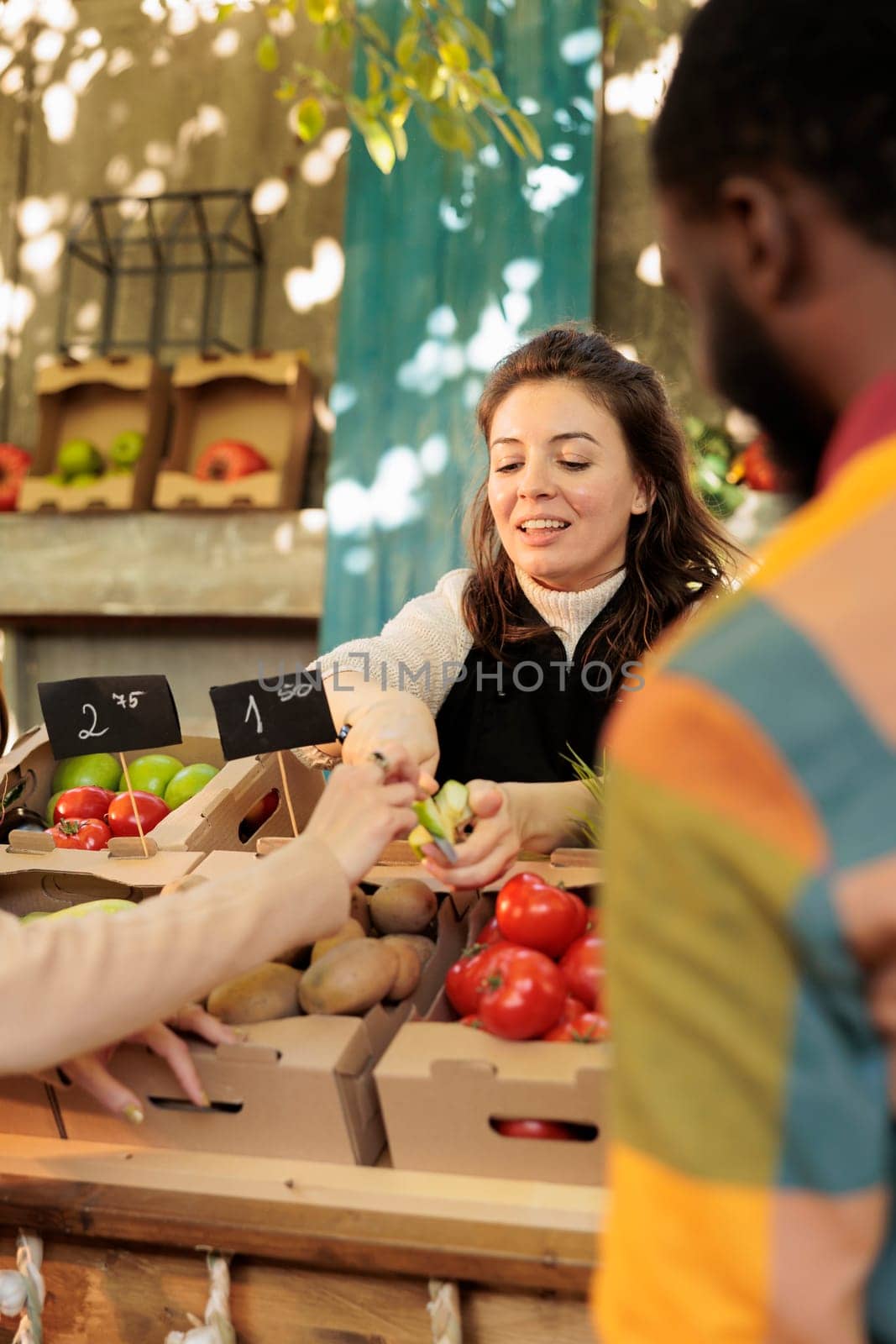 Smiling friendly organic fruits and vegetables shop owner offering customer to taste different varieties of apples at food market. Positive female farmer selling fresh local homegrown products