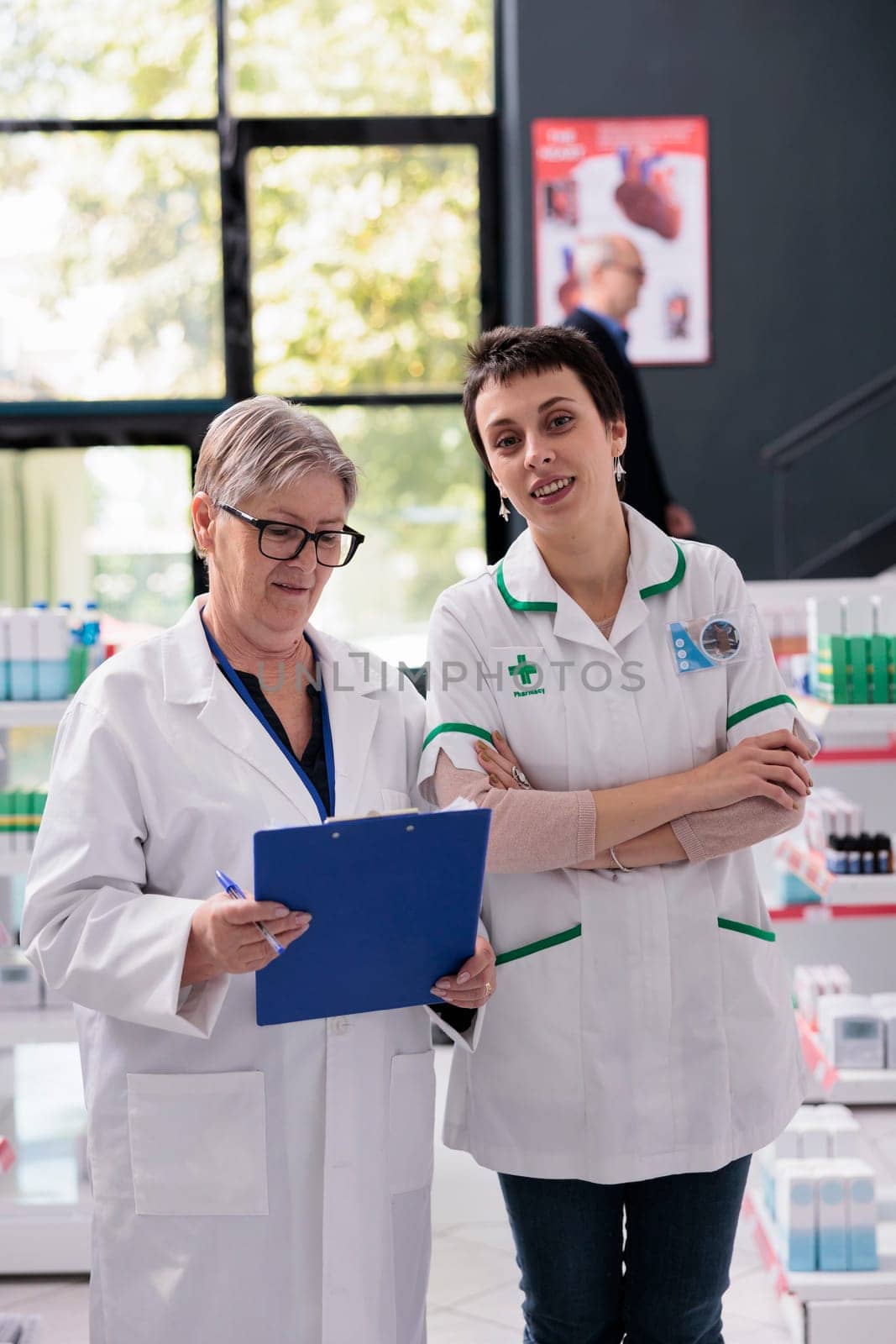 Two smiling woman pharmacy specialists in drugstore portrait. Elderly pharmaceutical employee holding prescription clipboard and young apothecary worker looking at camera