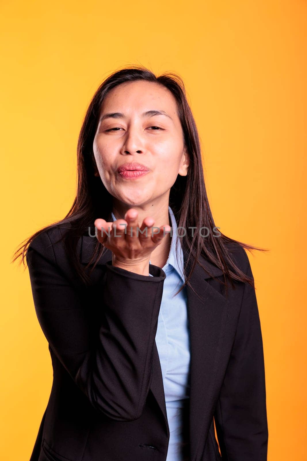 Attractive asian model blowing air kissed during studio shot, posing over yellow background. Confident carefree woman expressing love, looking at the camera with a satisfied smile. Romantic gesture