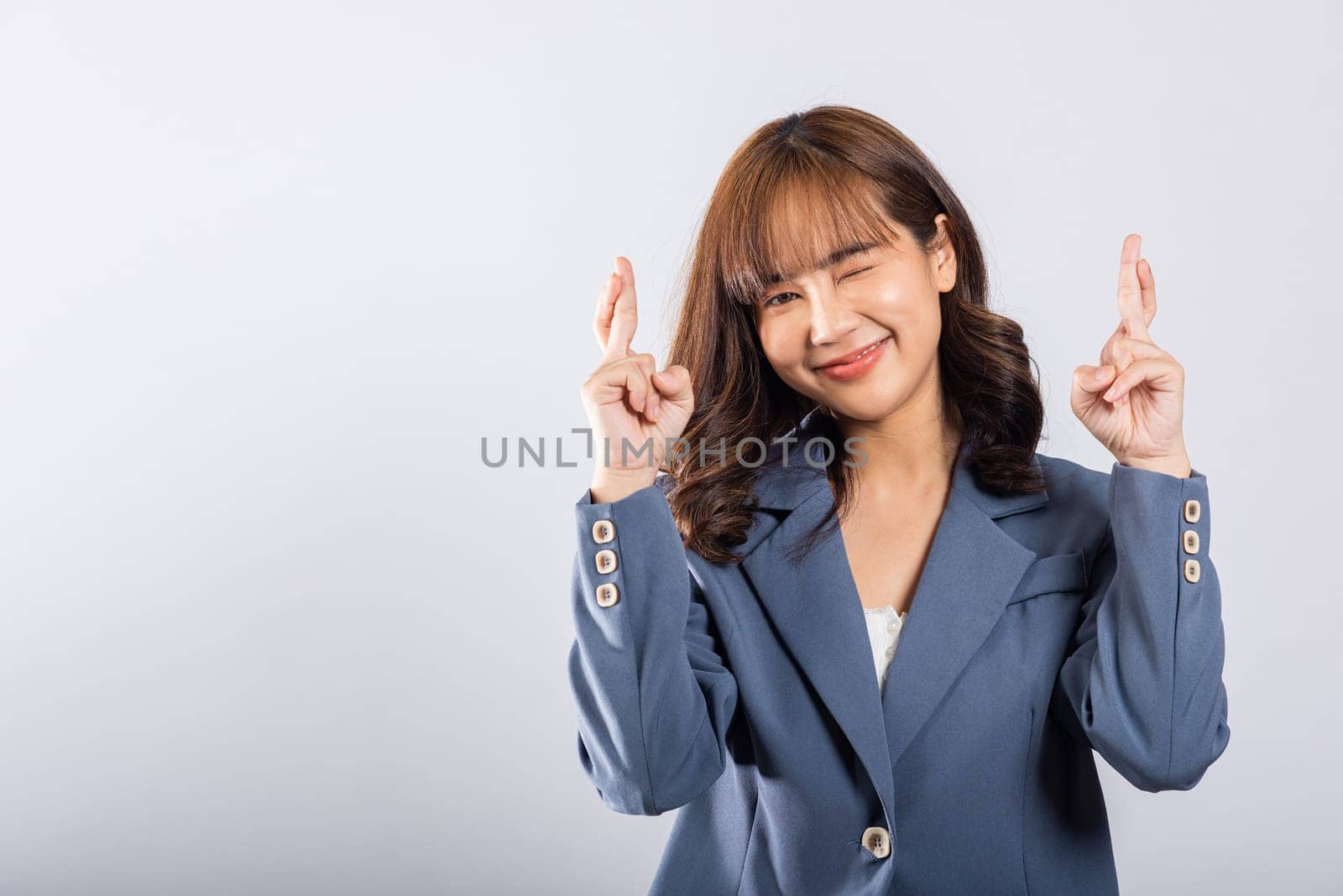A beautiful young Thai woman is captured in a studio shot, her smiling face reflecting happiness and hope. With crossed fingers, she silently conveys her belief in good luck and positive vibes.