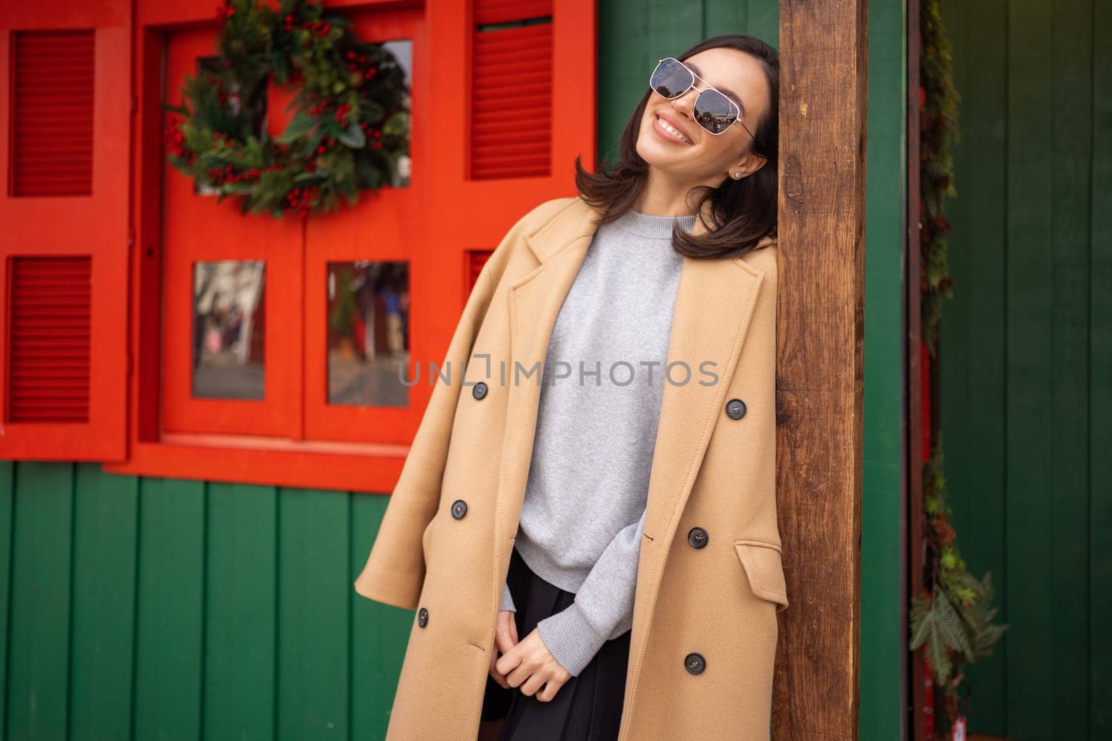 People, season and Christmas concept portrait of happy smiling middle age woman outdoors dressed stylish cream trench coat with Christmas festive decoration on background. Copy space