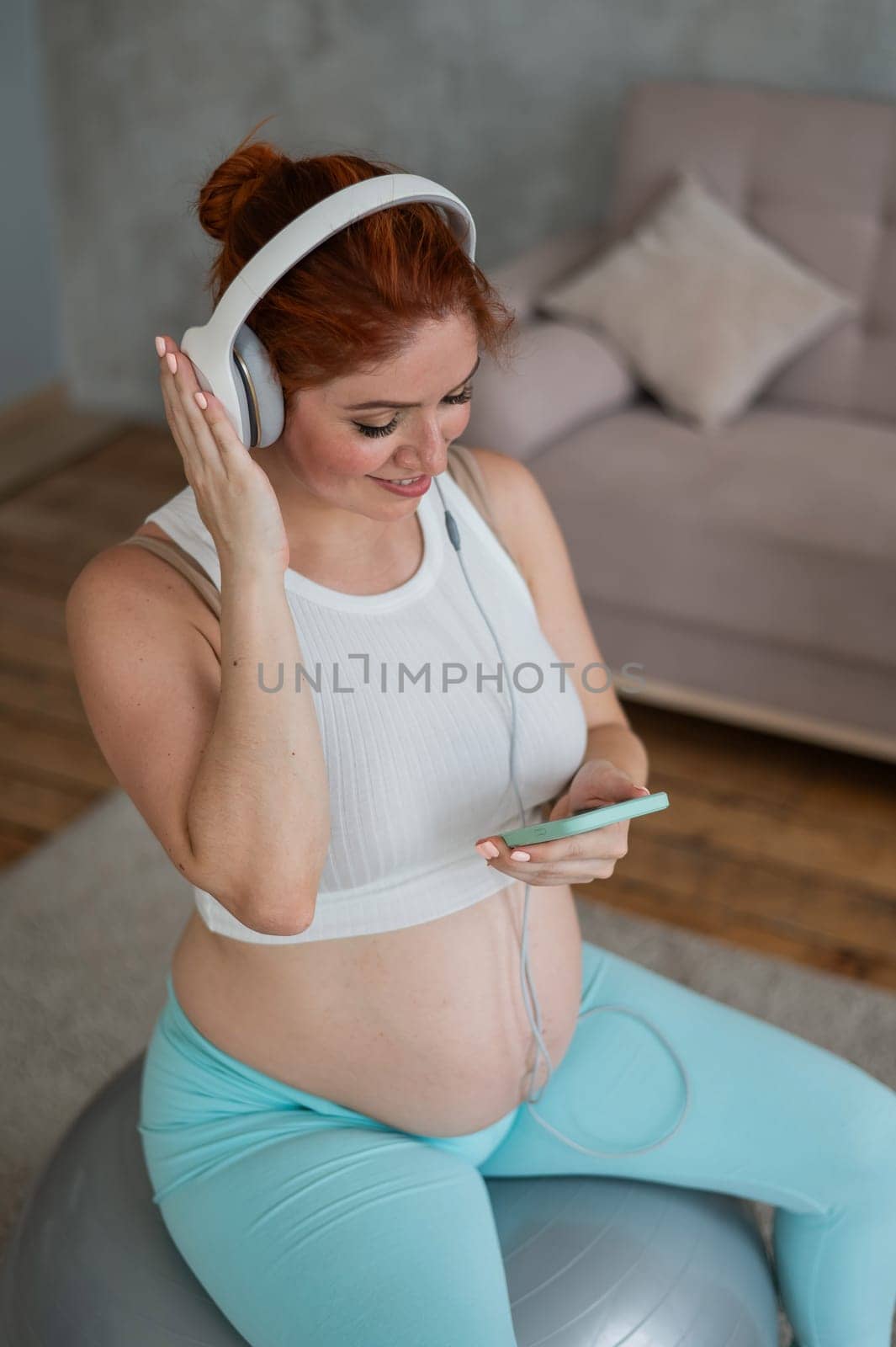 Caucasian red-haired pregnant woman sitting on fitball and listening to music with headphones at home. by mrwed54