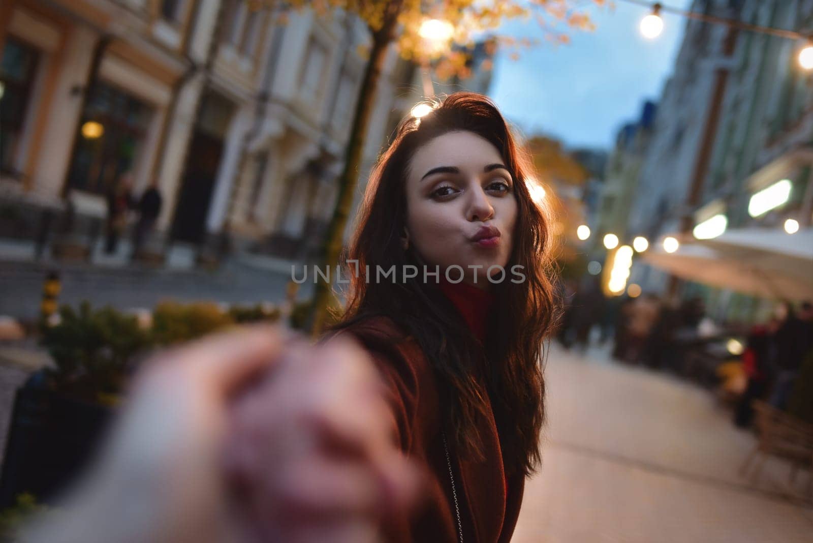 follow me. Beautiful romantic brunette girl in red coat takes her boyfriend's hands while walking in the evening city by Nickstock