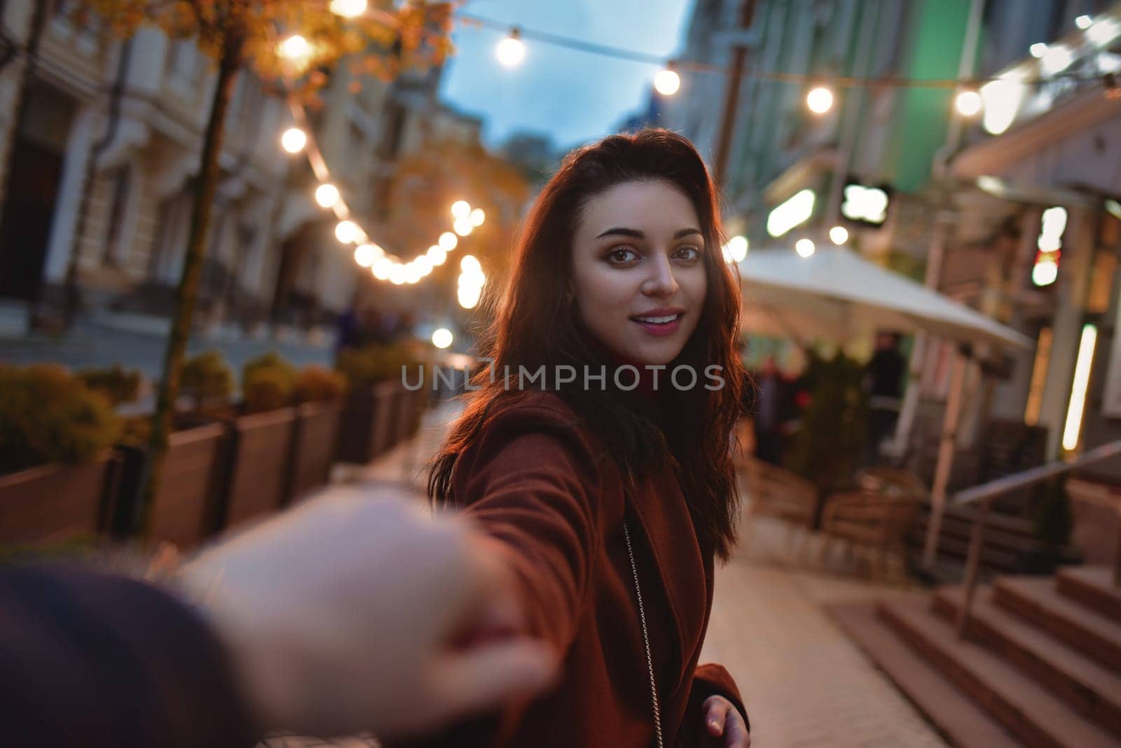 follow me. Beautiful romantic brunette girl in red coat takes her boyfriend's hands while walking in the evening city by Nickstock