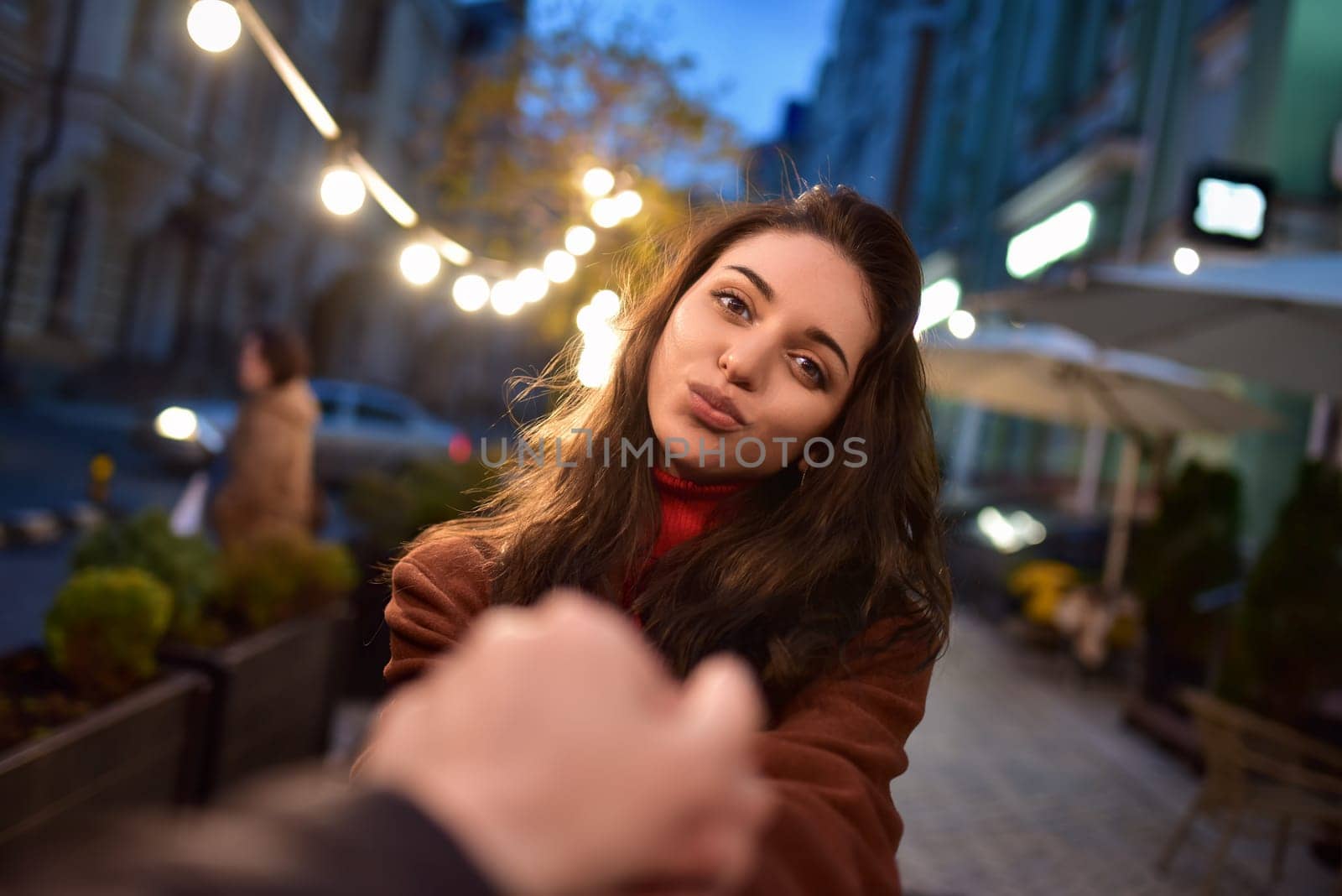 follow me. Beautiful romantic brunette girl in red coat takes her boyfriend's hands while walking in the evening city by Nickstock