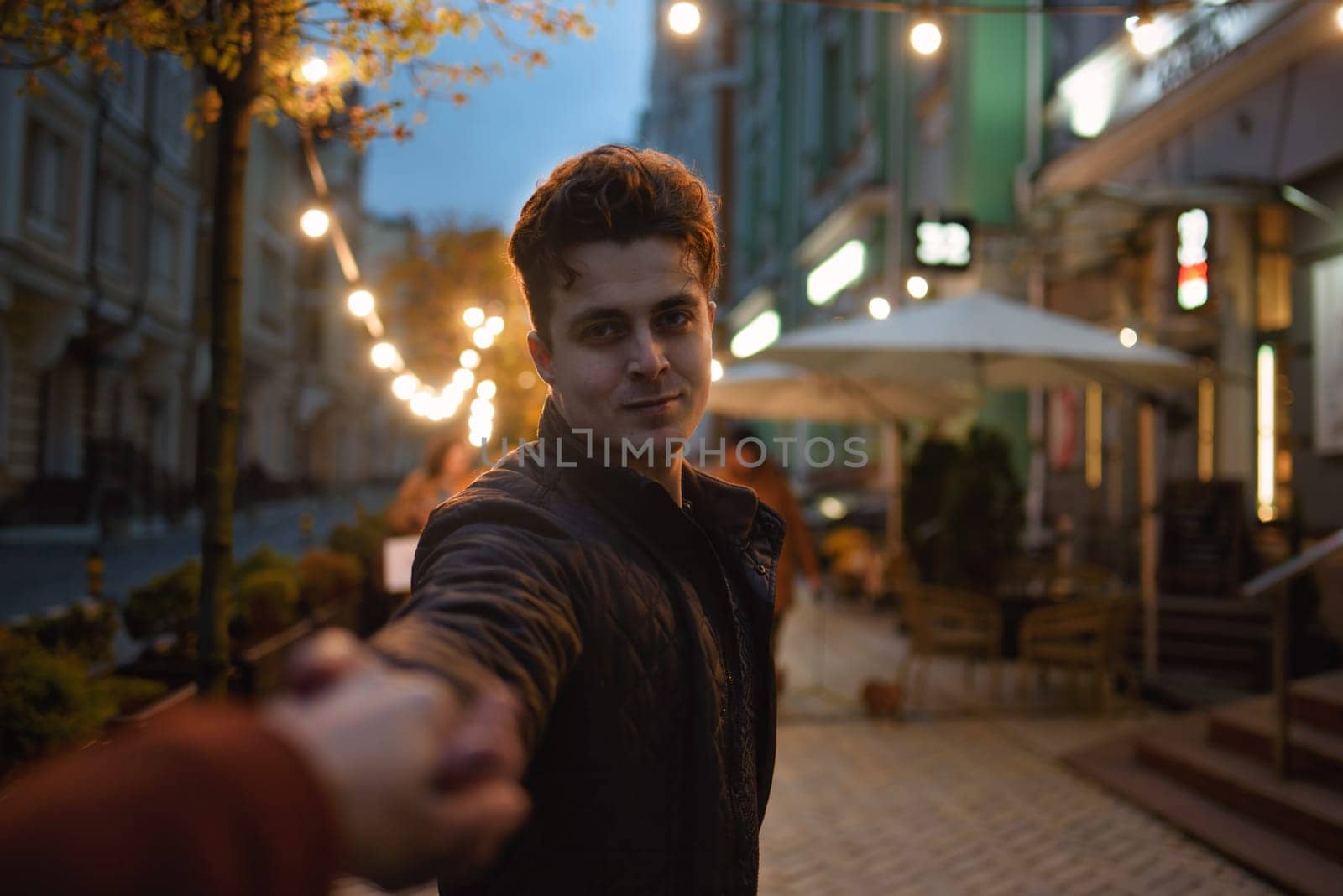 follow me. Beautiful romantic curly man takes his girlfriend hand while walking in the evening city by Nickstock