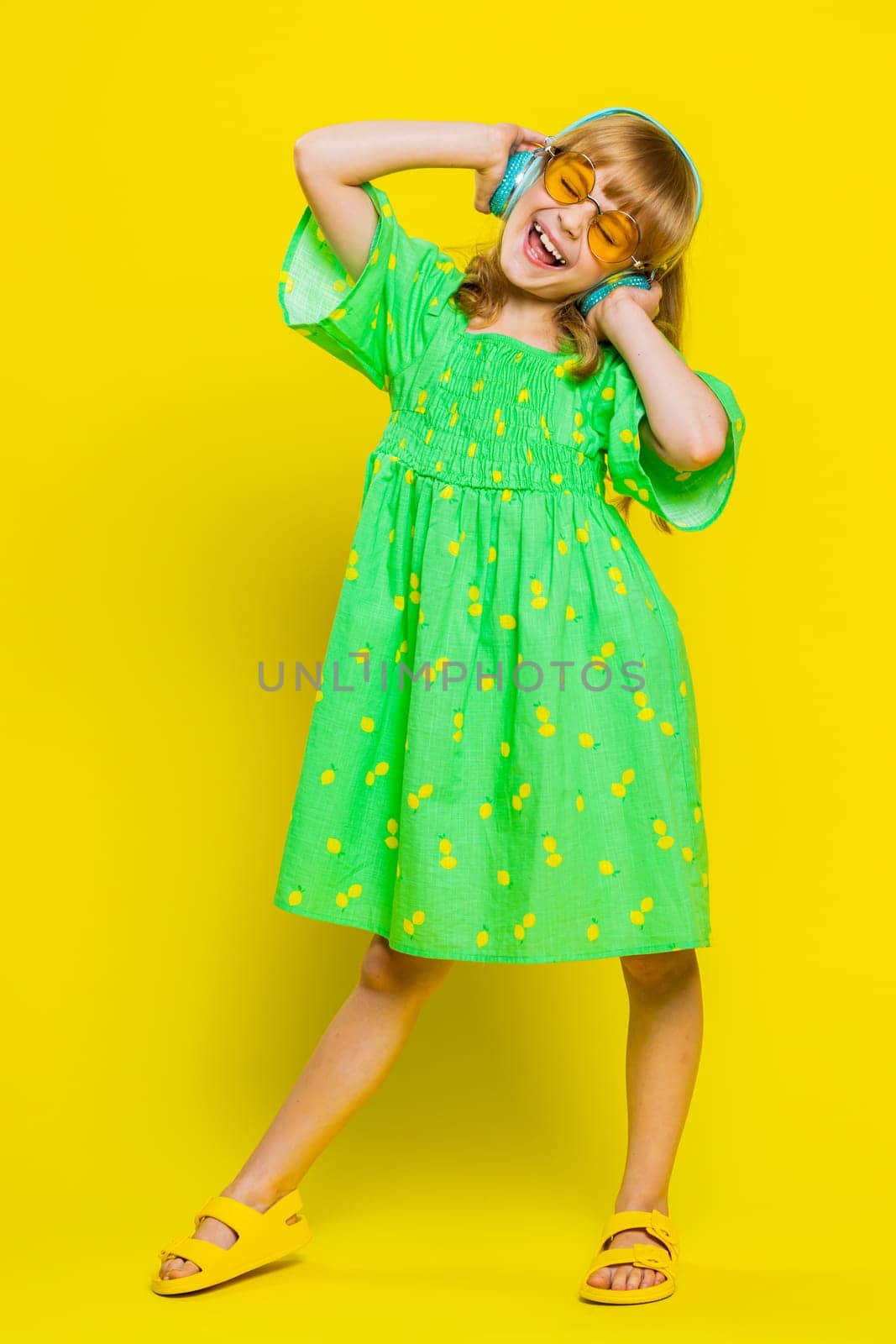 School girl listening music via headphones and dancing disco party fooling around having fun expressive gesticulating hands. Preteen Caucasian child kid in sunglasses on yellow background. Vertical